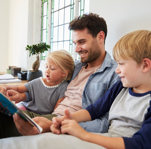 A man sits on a couch reading a book to two smiling young children, a girl and a boy, in a bright room with large windows, sharing quality time much like at a Greater Boston Fathers Family Network Meeting.