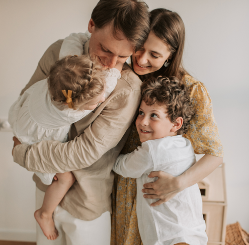 A family of four hugs and smiles warmly indoors, sharing a tender moment together. Their close bond highlights the power of family support, as they embrace with joy and warmth, radiating happiness and togetherness.