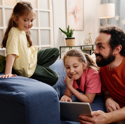 A man and two young girls sit on a blue couch at home, smiling and looking at a tablet together—enjoying moments of analog parenting in a digital world. The living room has a plant, warm lighting, and cozy decor in the background.
