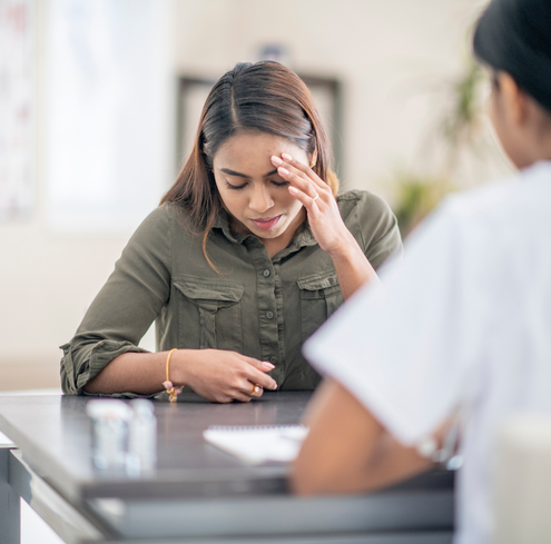 A woman sits at a desk with her head resting on one hand, looking stressed or worried, while another person, possibly a doctor or counselor, sits across from her with a notebook—highlighting concerns about parental mental health.