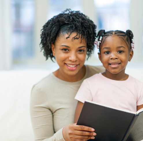 A smiling woman and a young girl sit together on a couch, holding an open book. Their shared moment of narrative self-expression fosters understanding, as stories bring them closer and help build community.