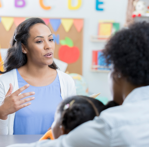 A teacher sits at a table, talking to a parent and child in a colorful, student-centered classroom, with educational decorations and alphabet letters visible in the background, highlighting the importance of family support.