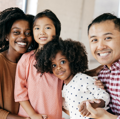 A group of four people, two adults and two children, stand close together indoors in Hampden County, smiling warmly at the camera. They appear happy and affectionate, showing the strong bonds of a true Family Network with arms around each other.