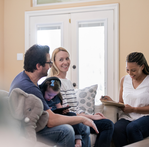 A smiling woman sits on a couch with a man and child, while another woman with a clipboard discusses national strategies to reduce unnecessary reports. The child holds a tablet, and the group enjoys a friendly discussion in a bright living room.