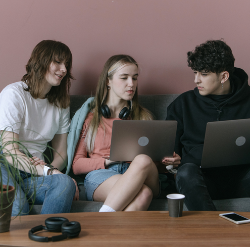 Three young people sit on a couch with laptops, engaged in conversation during a training session. One wears headphones around their neck, and a plant and headphones are on the table in front of them.
