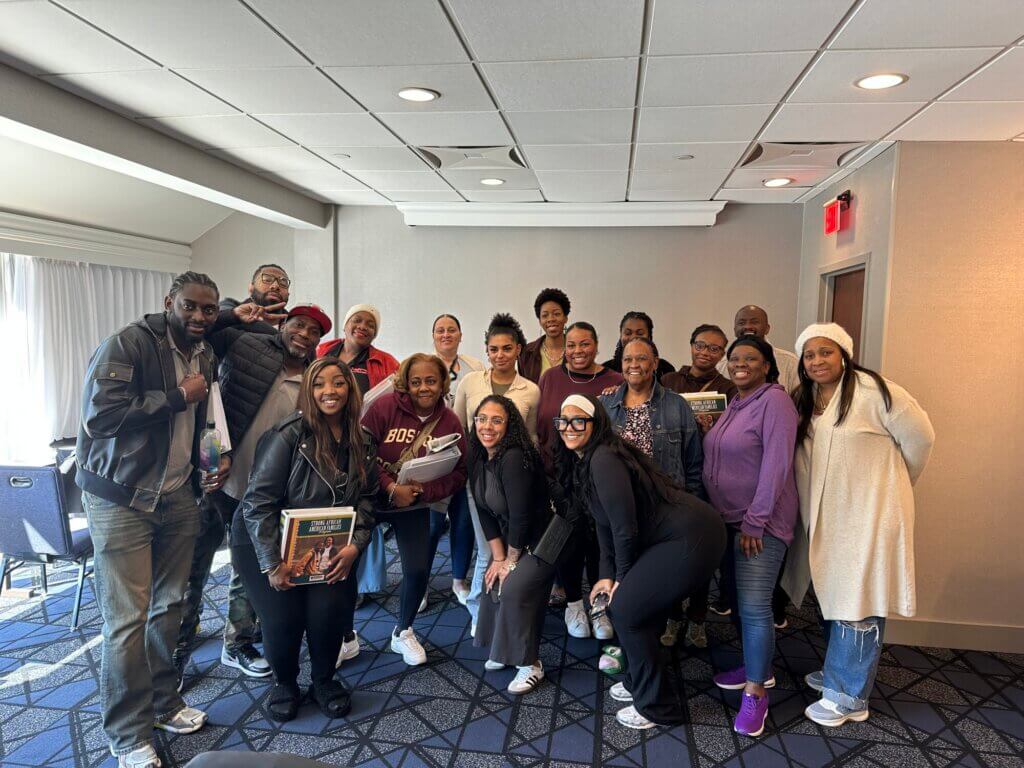 A group of about twenty people smile and pose together in a conference room with blue carpet and beige walls; some hold awards or folders, representing the Strong African American Families (SAAF) program, and everyone is dressed casually.
