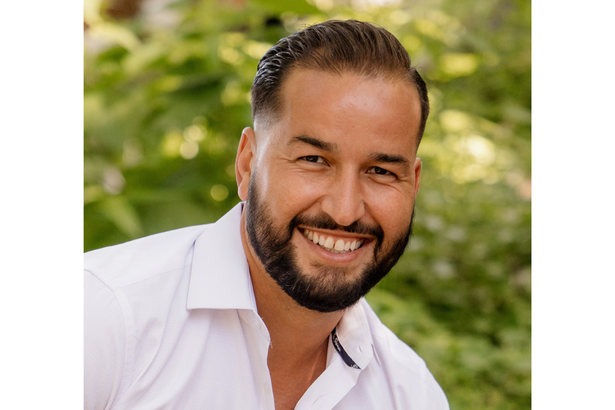 A man with dark hair and a beard, wearing a white collared shirt, smiles at the camera. The background is blurred greenery, suggesting an outdoor setting—perfect for a profile photo of Michaela Gagne, CAGS, LMHC.