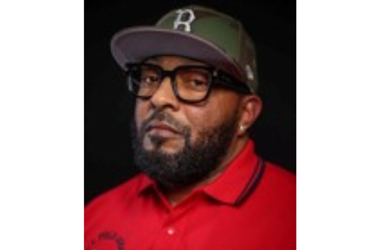 A bearded man wearing black-rimmed glasses, a camouflage baseball cap with a B on it, a red collared shirt, and a stud earring, poses against a dark background.