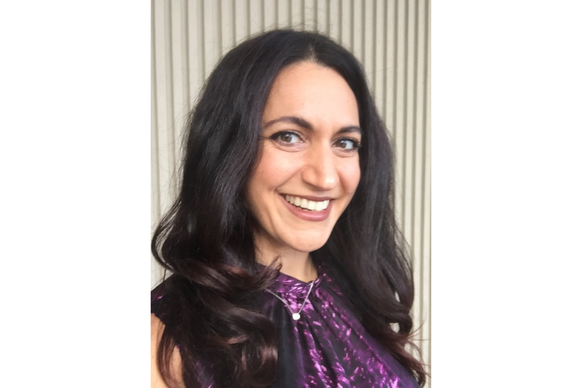 Lenore Maniaci, MPH, with long dark hair and a purple patterned top, smiles at the camera while standing in front of a beige vertical-striped background.