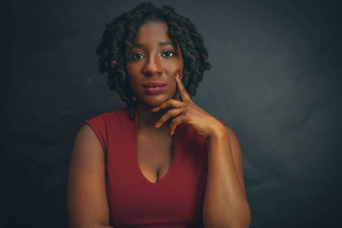 Jennifer Byrd, a woman with short curly hair in a sleeveless maroon top, poses against a dark background, resting her chin on her hand and looking thoughtfully at the camera.