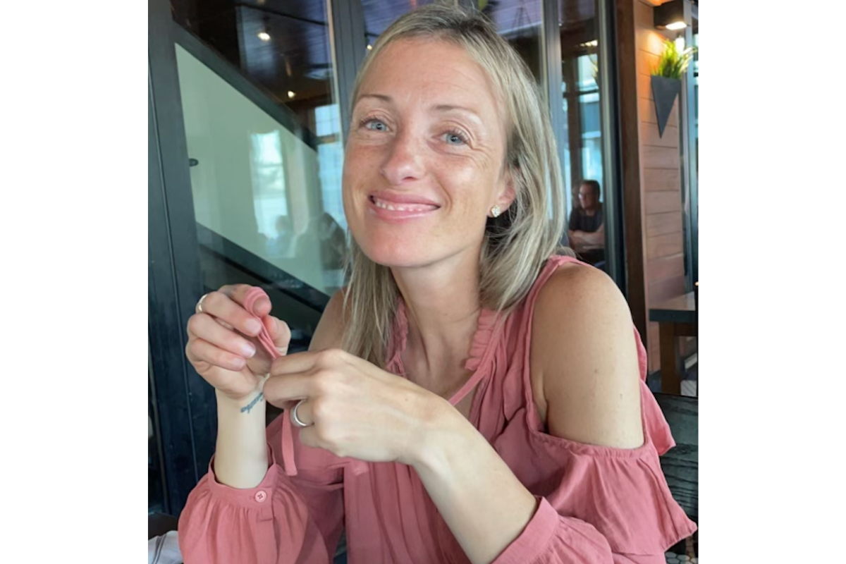 Alexis Florence, with long blond hair and a pink cold-shoulder blouse, sits indoors at a table, smiling at the camera while holding a tea bag string in her hands.