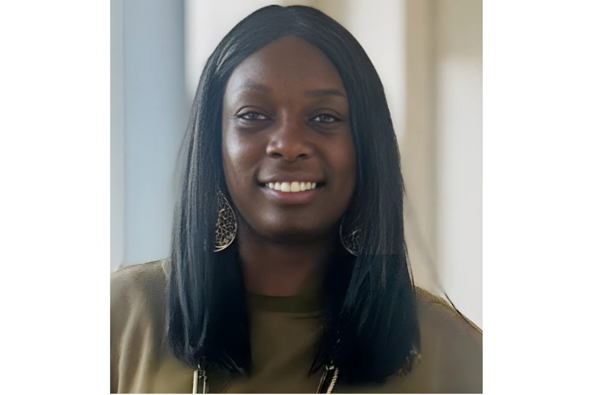 T'Sheba Martin, a woman with long, straight black hair, is smiling. She is wearing a green top, large dangling earrings, and a necklace. The background is softly blurred with natural light coming through a window.