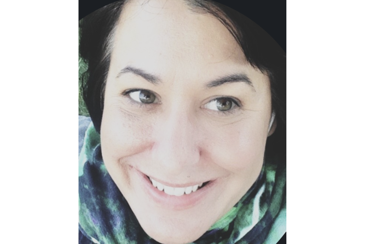 A close-up photo of Kirsten Durzy, MPH, a person with light skin, dark hair, and hazel eyes, smiling and wearing a green and blue patterned scarf. The background is out of focus.