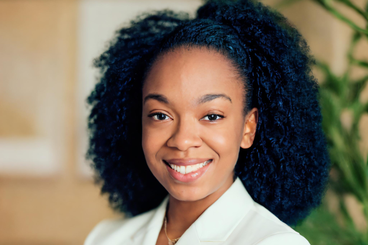 LaTasha Reece, a woman with natural curly hair, smiles warmly in a light-colored blazer, standing indoors with a softly blurred background of neutral tones and green plant leaves.