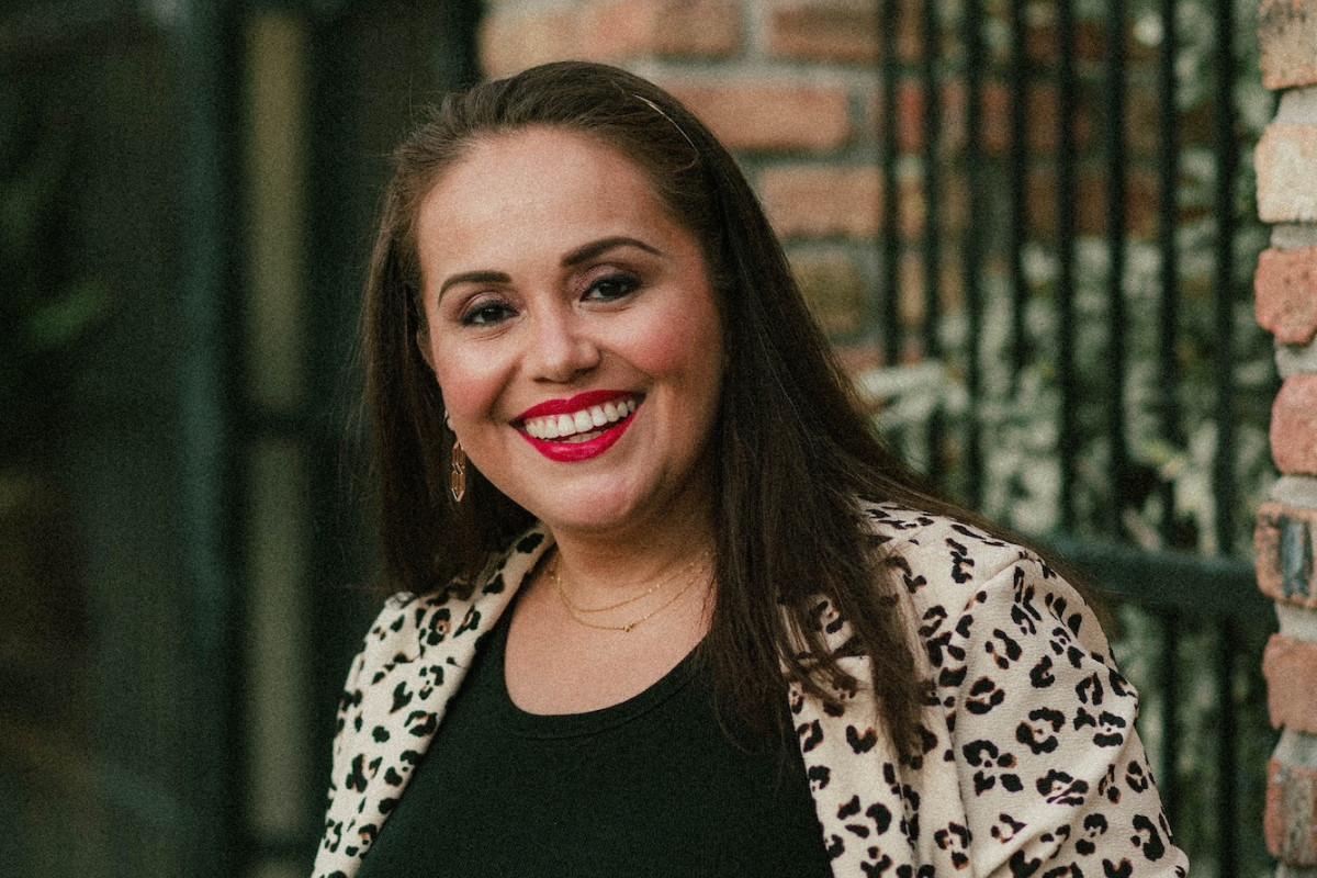 Christina Kozik, with long brown hair, smiles warmly in a leopard print jacket over a black top. Her bright red lipstick and gold jewelry stand out as she poses in front of a brick wall with a metal fence.