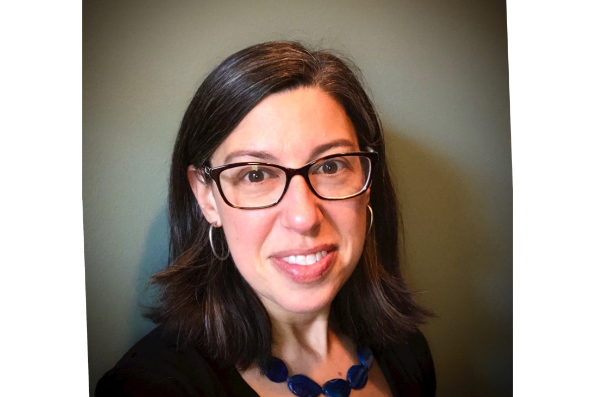 Marisa Howard-Karp, a woman with shoulder-length dark hair and glasses, smiles at the camera. She is wearing a black top, hoop earrings, and a chunky blue necklace, with a plain greenish background behind her.