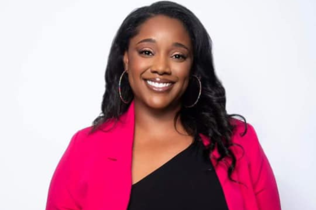 Krystina Jackson, MS, with long dark hair, smiles at the camera, wearing hoop earrings, a bright pink blazer, and a black top as she stands against a plain white background.