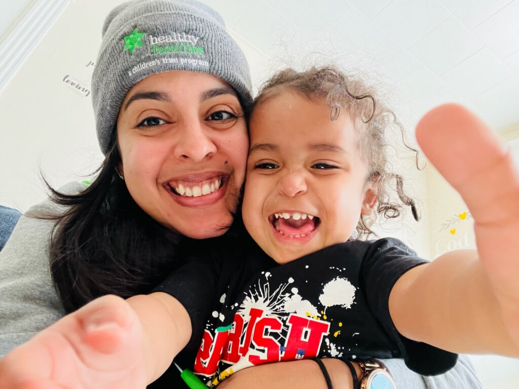 A smiling woman in a gray beanie and a cheerful young child with curly hair take a close-up selfie together, both reaching toward the camera, appearing happy and playful as they celebrate National Home Visiting Week.