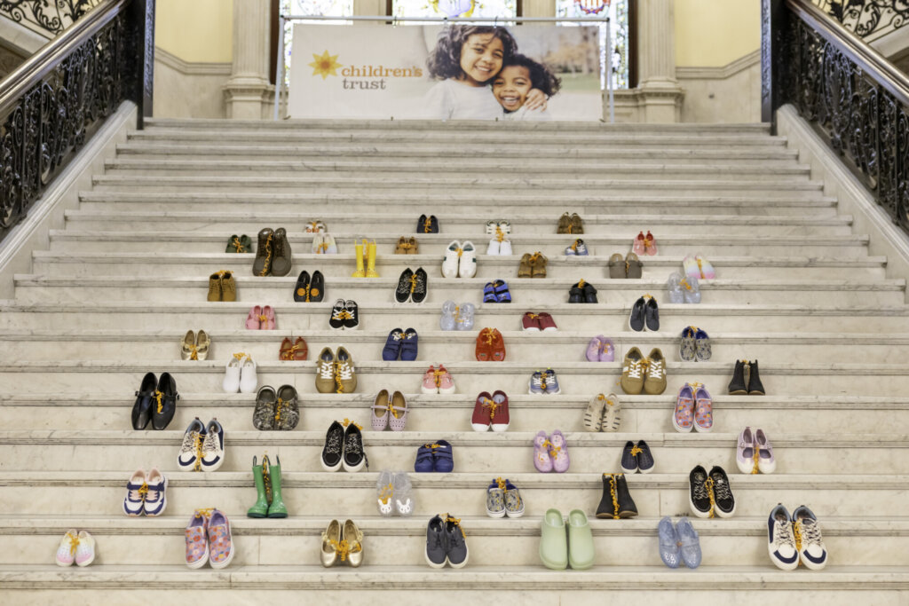 Pairs of children’s shoes arranged neatly on marble steps at the Massachusetts State House, with a large banner above showing two children smiling and the words “children’s trust.” Ornate railings frame this Stepping Up for Kids advocacy display.