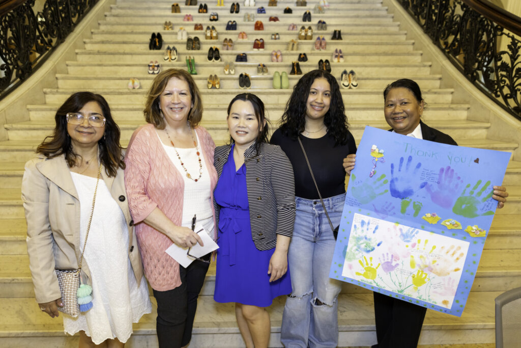 Five women stand smiling on marble stairs lined with shoes at the Massachusetts State House. One holds a colorful “Thank you” poster, celebrating Stepping Up for Kids and their dedication to kids advocacy in a formal setting with ornate railings.