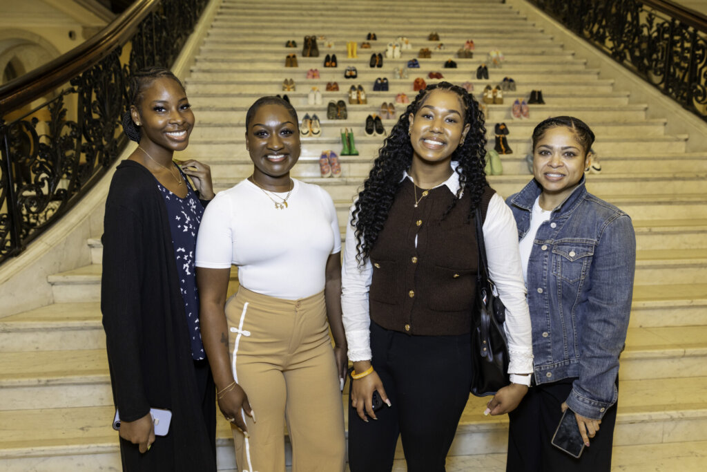 Four women smiling and posing together in front of a grand staircase at the Massachusetts State House, decorated with many pairs of colorful kids' shoes arranged in rows for Stepping Up for Kids.