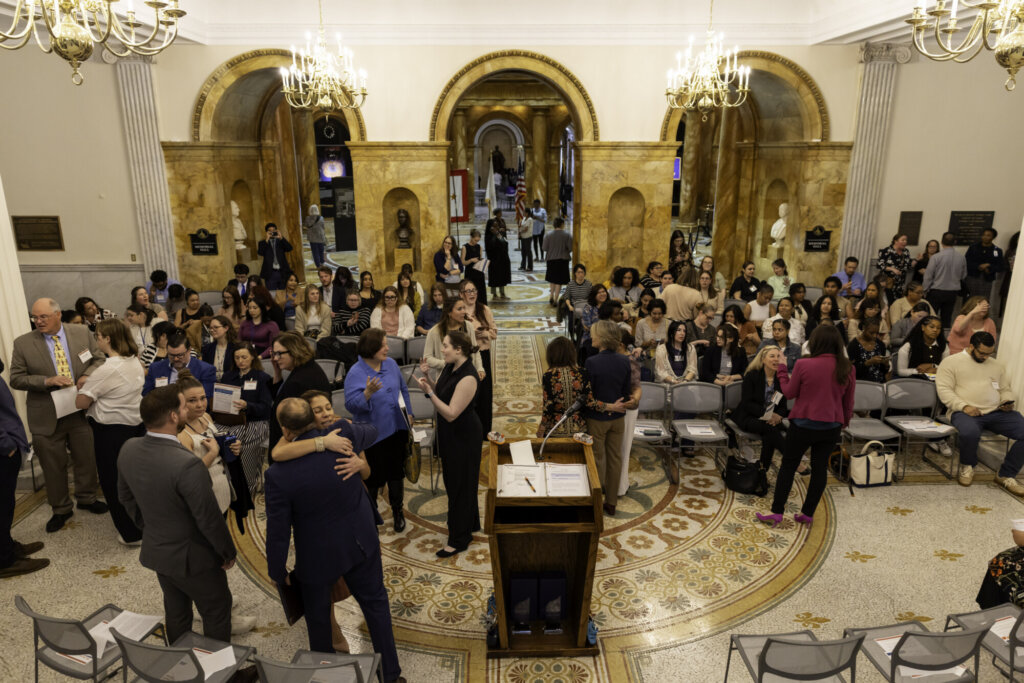 A large group gathers in an ornate hall at the Massachusetts State House, with chandeliers, marble columns, and patterned flooring. Attendees talk, hug, and prepare for the Stepping Up for Kids event amid rows of chairs.