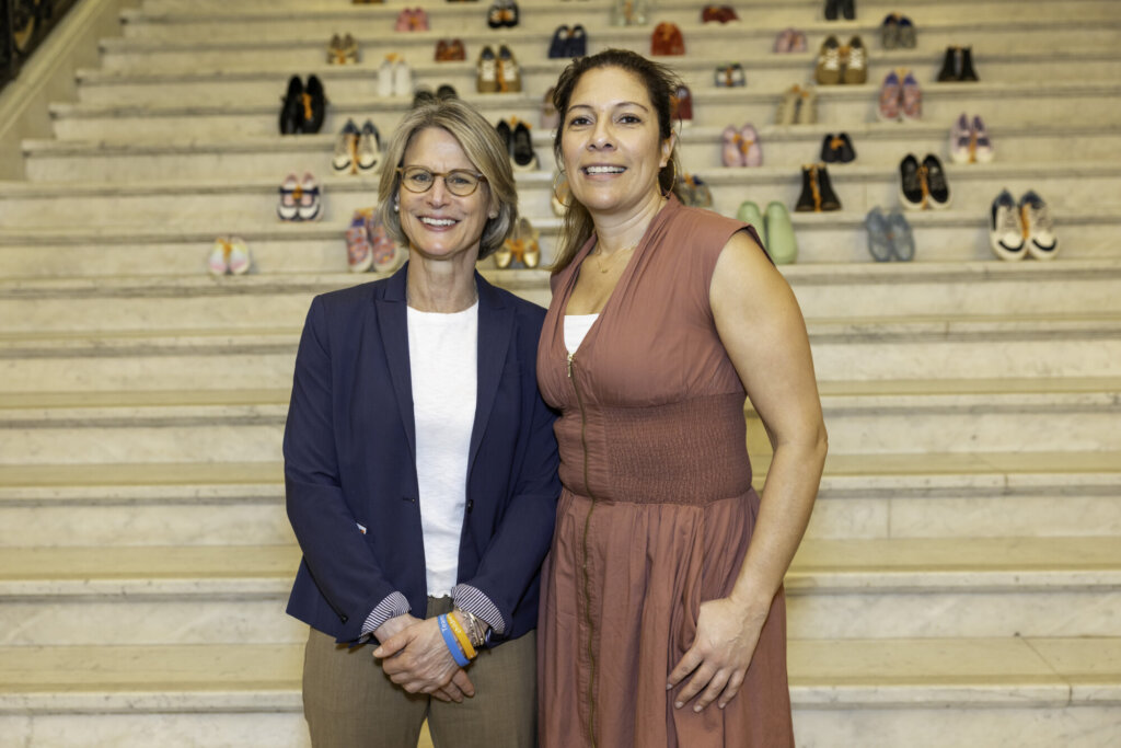 Two women stand side by side, smiling, in front of the Massachusetts State House’s marble stairs, where pairs of children's shoes are arranged in rows as part of the Stepping Up for Kids display.