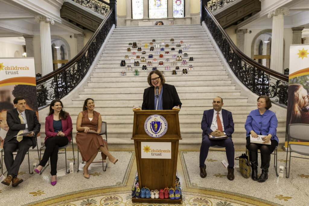 A woman speaks at a podium in the ornate Massachusetts State House, stepping up for kids amid a grand staircase lined with children’s shoes. Five people listen as banners display the Children’s Trust logo, highlighting powerful kids advocacy.