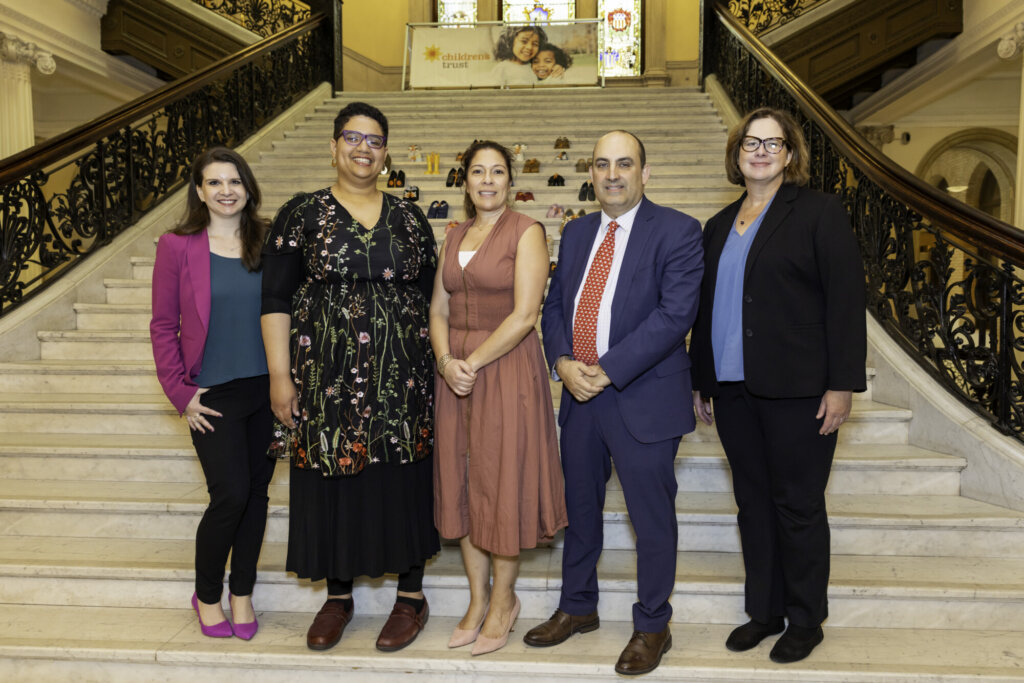 Five people in business attire stand on marble stairs with ornate railings at the Massachusetts State House. Behind them are pairs of small shoes and a banner with children’s faces, highlighting their commitment to kids advocacy.