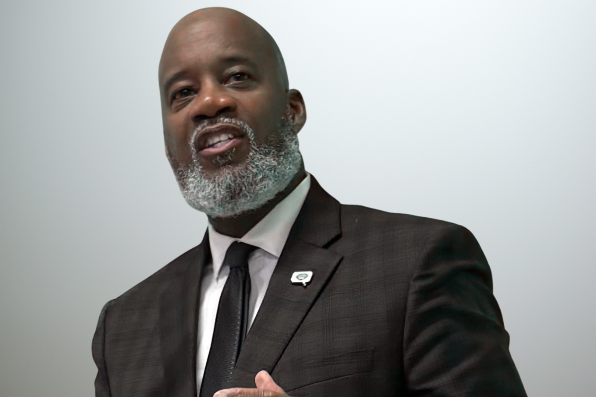 Abdul-Rahmaan Muhammad, a man in a dark suit and tie with a gray beard, is speaking and gesturing with his hand. He wears a pin on his lapel and stands against a light, plain background.