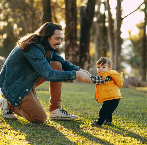A man in a denim jacket kneels on grass, holding hands with a toddler in a yellow vest as they stand outdoors in a sunlit, wooded park, capturing the warmth of fathers influence on early literacy.