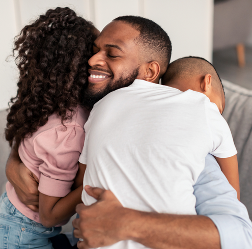 A smiling father with a beard hugs two children closely in a bright room, expressing warmth and affection. This moment captures the spirit of the Greater Boston Family Network.