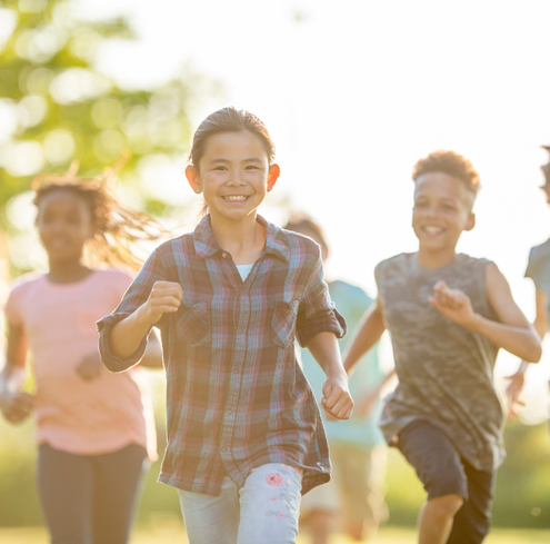 A group of smiling children run outdoors on a sunny day, with one child in a plaid shirt leading the group as if guiding a playful training session. The bright background is blurred with lush green foliage.