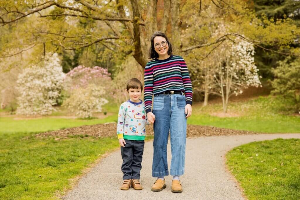 A smiling woman and young boy stand hand in hand on a park path, surrounded by spring blossoms