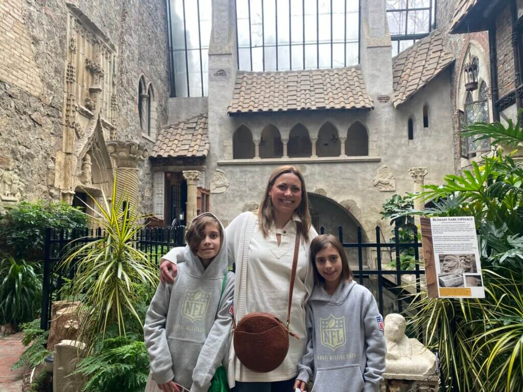 A woman stands smiling with two children in gray NFL hoodies inside a building with stone architecture, plants, and statues. The setting resembles a historic courtyard or museum exhibit, making for a memorable Mother's Day outing.