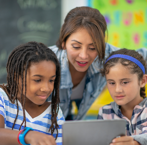 A teacher and two children are gathered around a tablet, smiling as they engage in training activities in a classroom filled with colorful decorations and a chalkboard in the background.