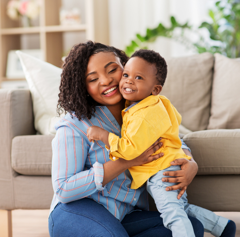 A smiling mother sits on the floor in a living room, hugging a cheerful young boy in a yellow shirt. They both look happy, and the background shows a couch and houseplants.