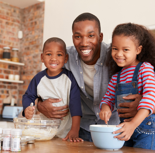 A smiling father stands between two young children as they mix ingredients in bowls at a kitchen counter, highlighting the joy of baking together and the strong family network they share.