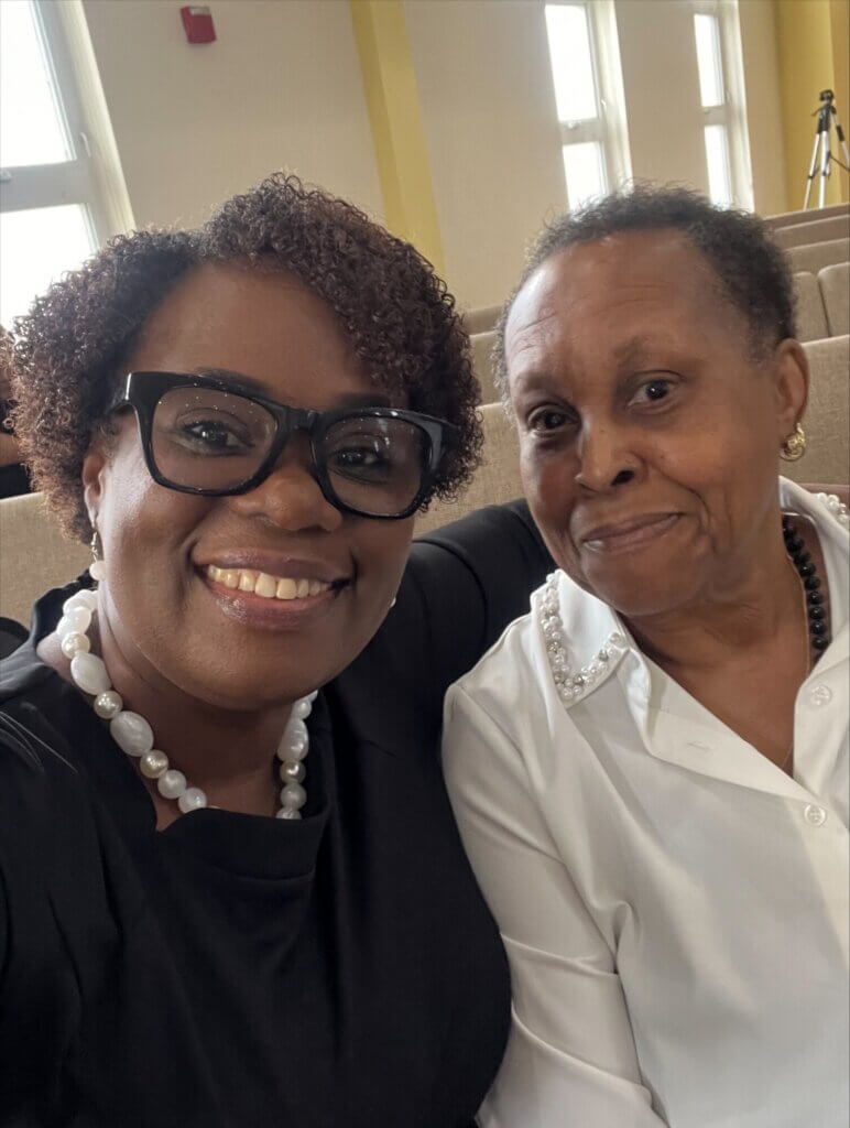 Two women smiling and sitting close together on beige seats indoors, celebrating Mother's Day—one wearing glasses and a black top with a pearl necklace, the other in a white blouse with pearl details. Bright windows shine in the background.