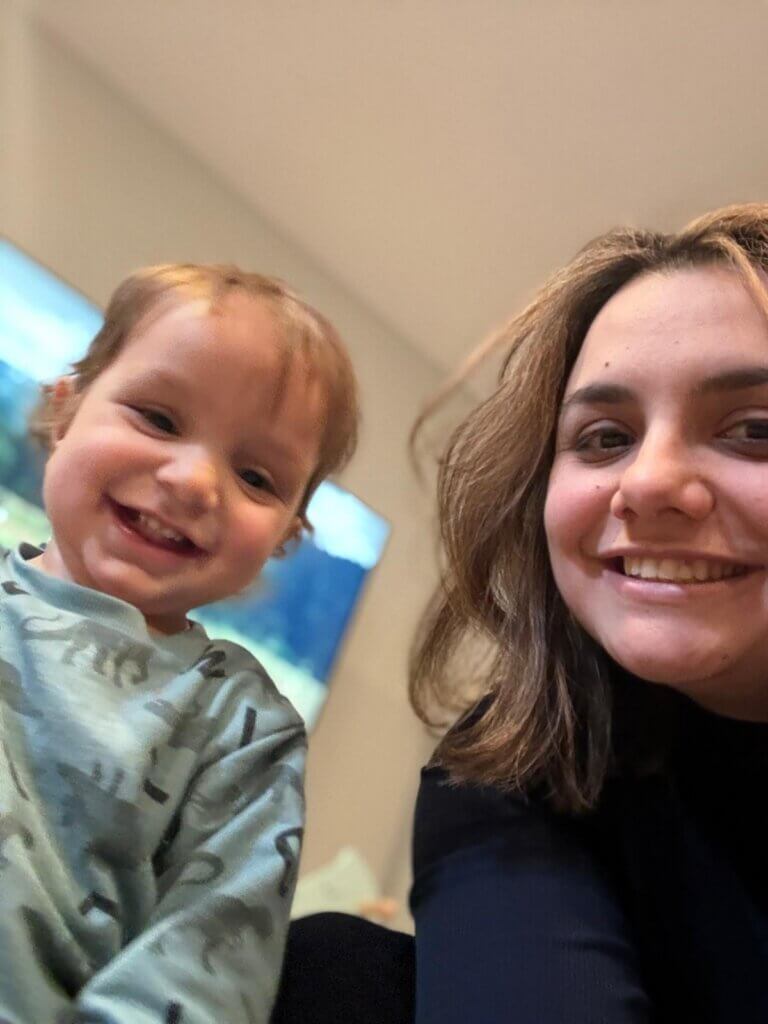 A smiling toddler in a light blue shirt with animal prints sits next to a woman with shoulder-length brown hair, both looking at the camera and smiling indoors, capturing a joyful Mother's Day moment that celebrates Children’s Trust.