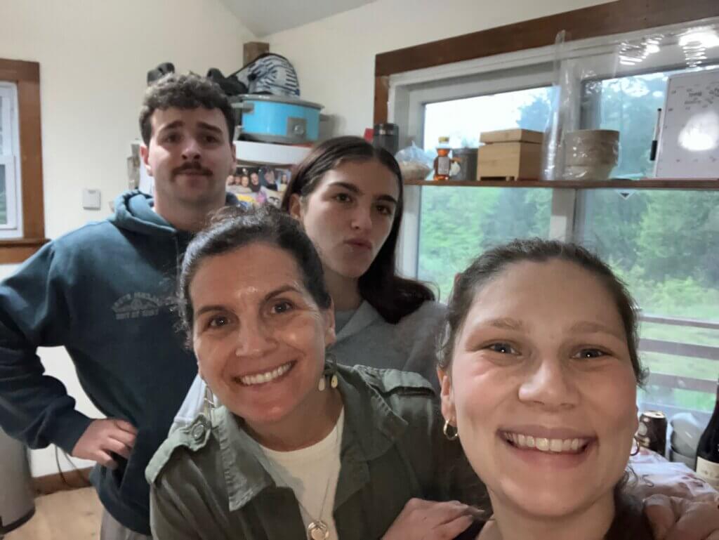 Four people stand close together smiling in a cozy kitchen. Two women are in the foreground, celebrating Mother's Day, while a young man and another woman stand behind them. A window shows greenery outside.