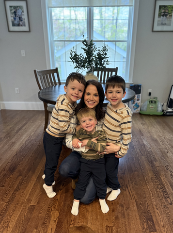 A woman kneels on a wooden floor, smiling and hugging three young boys in striped sweaters and white socks—capturing a joyful Mother's Day moment that radiates love and Children's Trust in a bright room with a round table and large window.