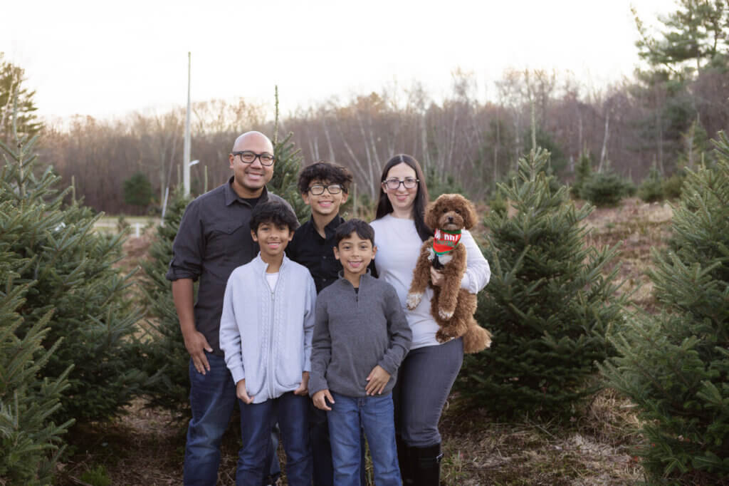 A family of five and a brown dog stand together outdoors among evergreen trees, smiling at the camera on what could be Mother's Day. The setting appears to be a Christmas tree farm in late autumn or early winter.