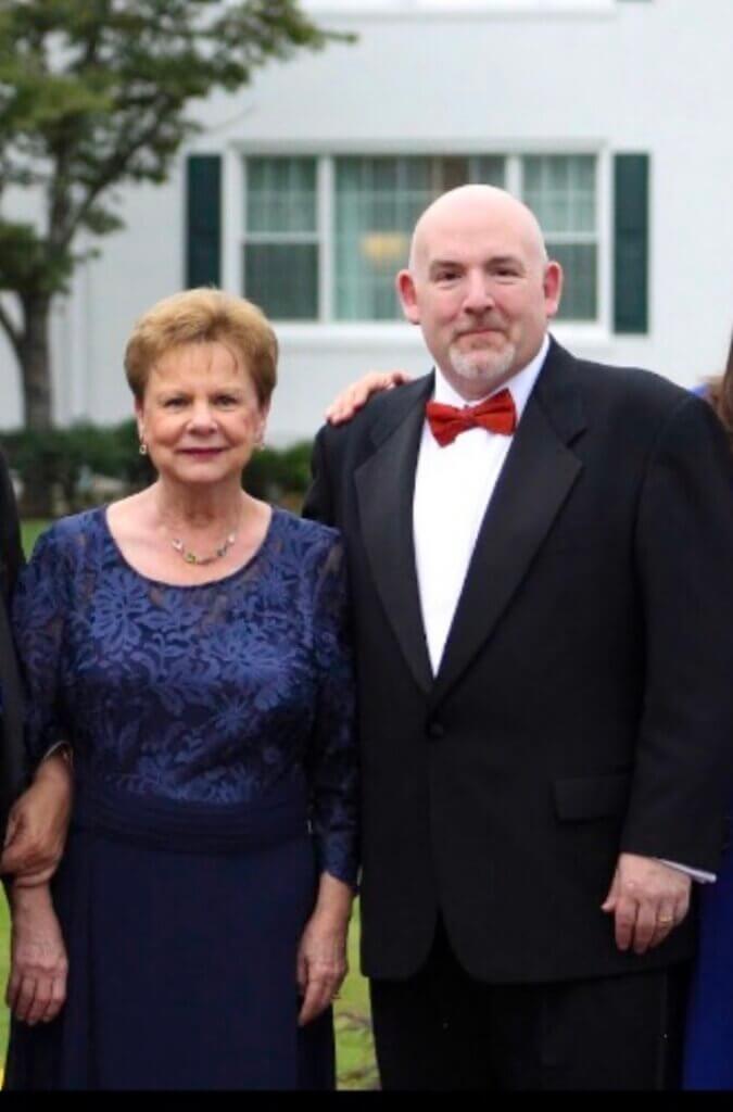 An older woman in a blue dress stands next to a man in a black tuxedo with a red bow tie. They are outdoors, smiling on Mother’s Day, with green trees and a white building in the background.