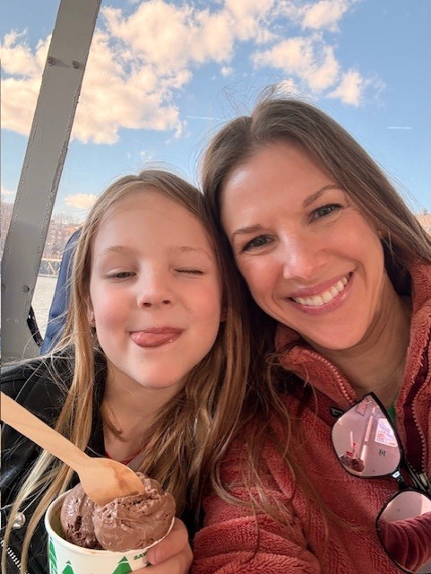 A woman and a girl smile for a selfie outdoors on Mother's Day, with the girl playfully sticking out her tongue and holding a cup of chocolate ice cream under a blue sky with scattered clouds. Their bond shines with true Children's Trust.