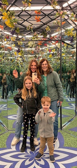 Two women and two children pose and smile for a Mother’s Day selfie in a mirrored room decorated with hanging flowers and a green and purple patterned floor. Their reflections create a kaleidoscopic effect, capturing joy and togetherness.