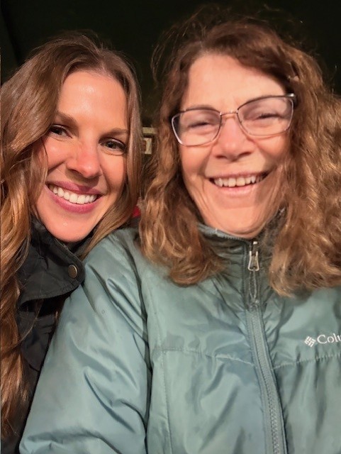 Two women smiling for a selfie—perfect for Mother's Day. The woman on the left has long brown hair and a dark jacket, while the woman on the right wears glasses, wavy hair, and a light green Columbia jacket—a moment of joy captured by Children's Trust.