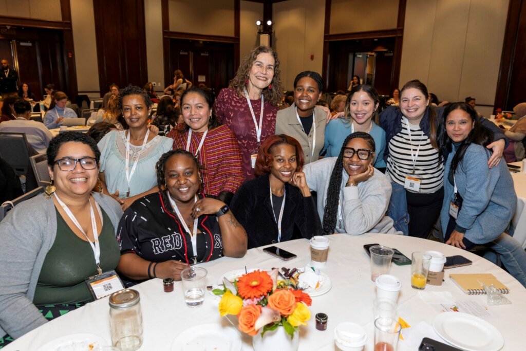 A diverse group of twelve smiling people pose together around a round table with drinks and a floral centerpiece at a A View from All Sides conference in a well-lit room, coming together.