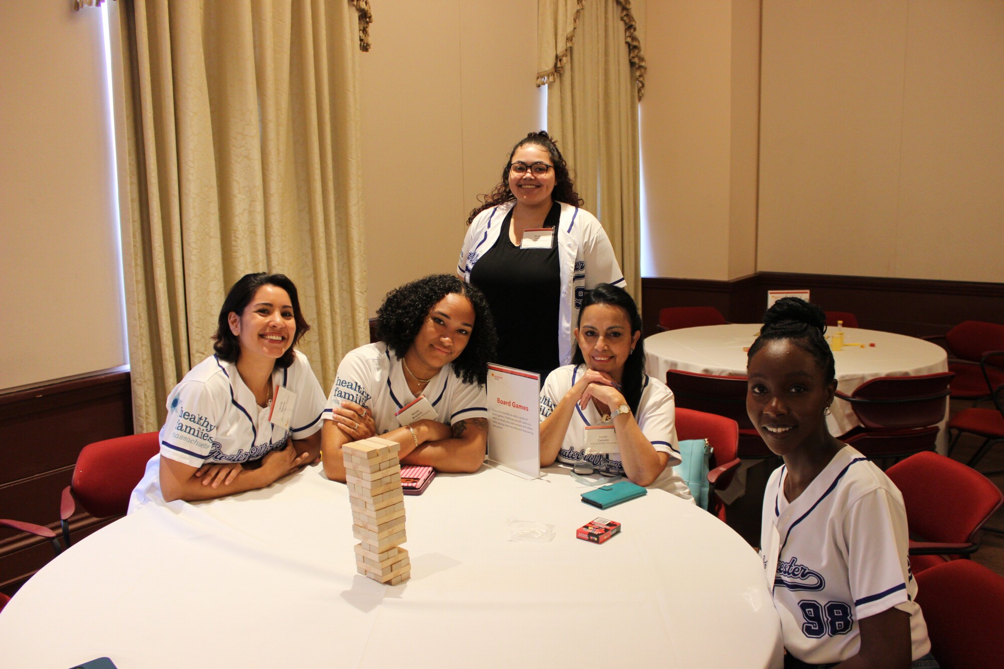 Five women sit and stand around a round table, smiling at the camera in matching white jerseys. A Jenga tower and game cards are on the table, capturing a Celebration Day honoring Massachusetts families in a conference room setting.