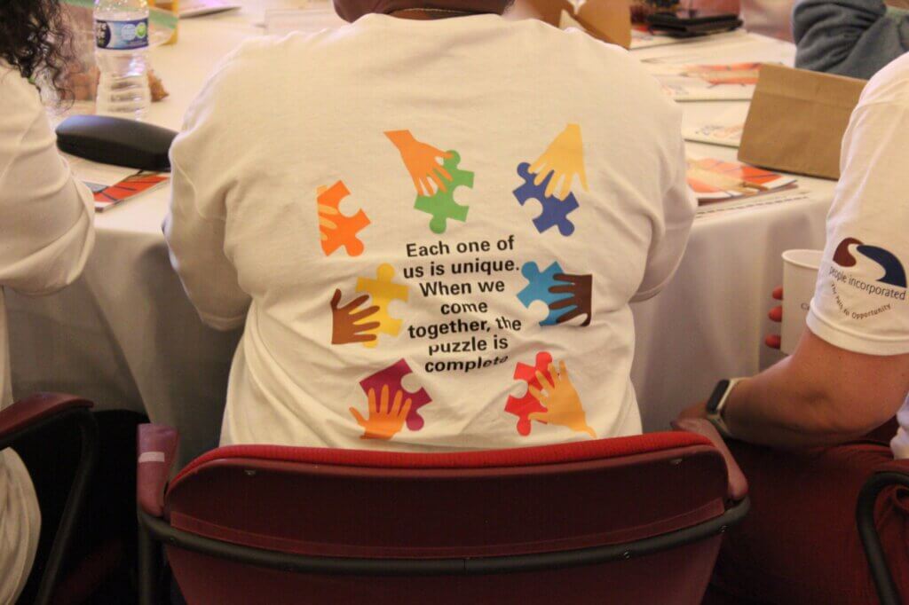 A person wearing a white shirt with colorful puzzle pieces and hands on the back, along with the text: Each one of us is unique. Honoring Massachusetts families, they are seated at a table with others, showing how together, the puzzle is complete.